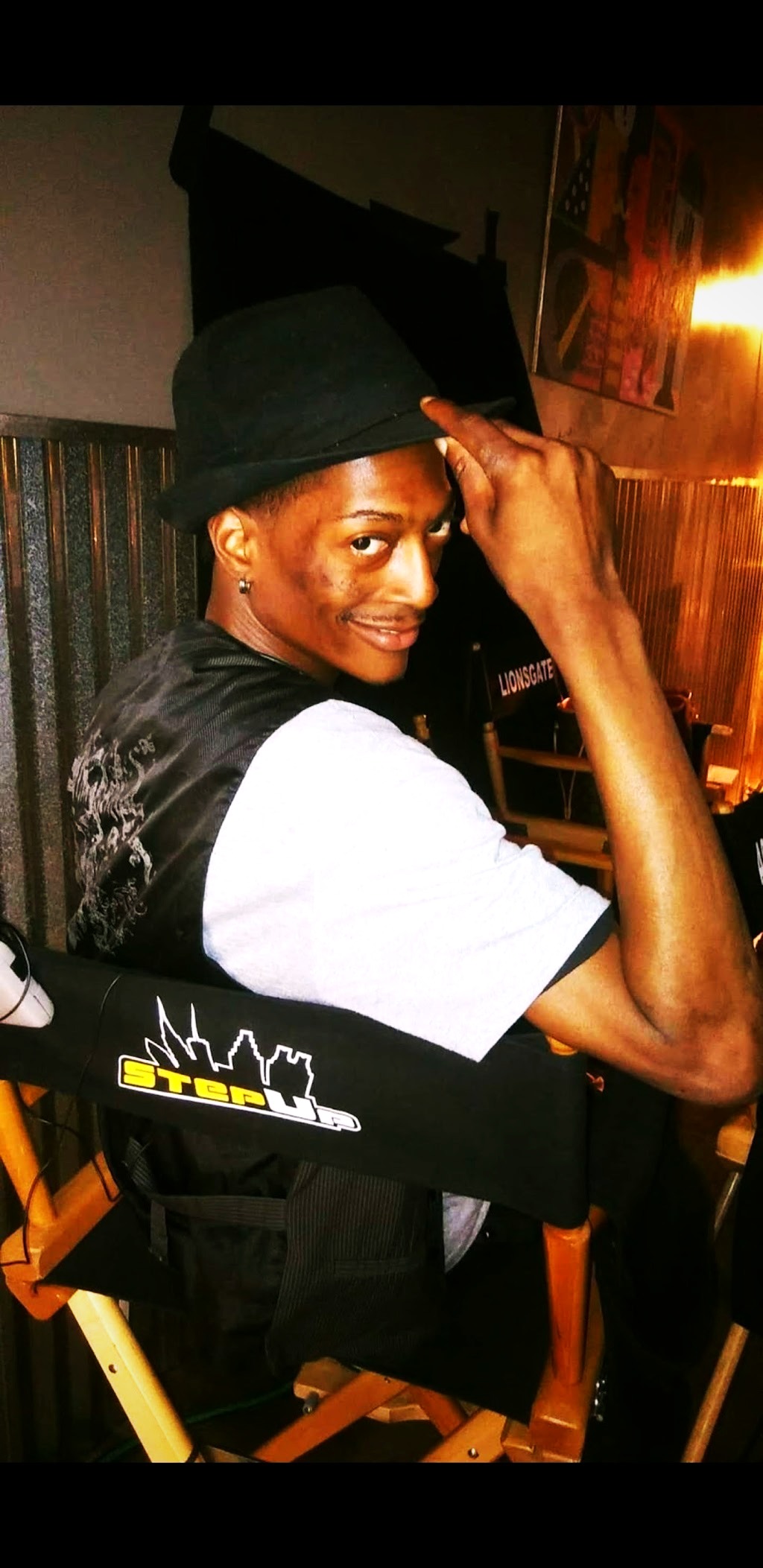 RikkiTheFool seated in a Step Up director's chair and tipping a black fedora behind the scenes